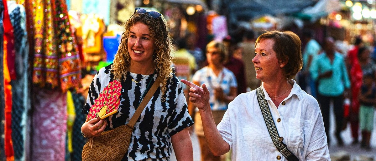 Two women walk through a bustling market street, one smiling and holding a red fan.