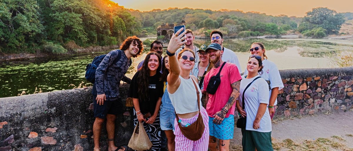 A group of travellers taking a selfie on a stone wall beside a lake at sunset.