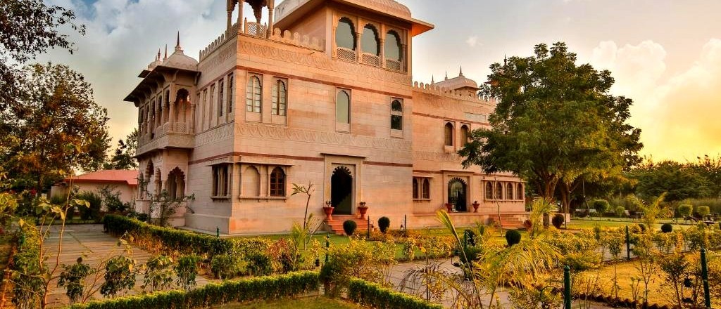 An ornate, light-coloured building with domes and arched windows stands in a lush garden at sunset.