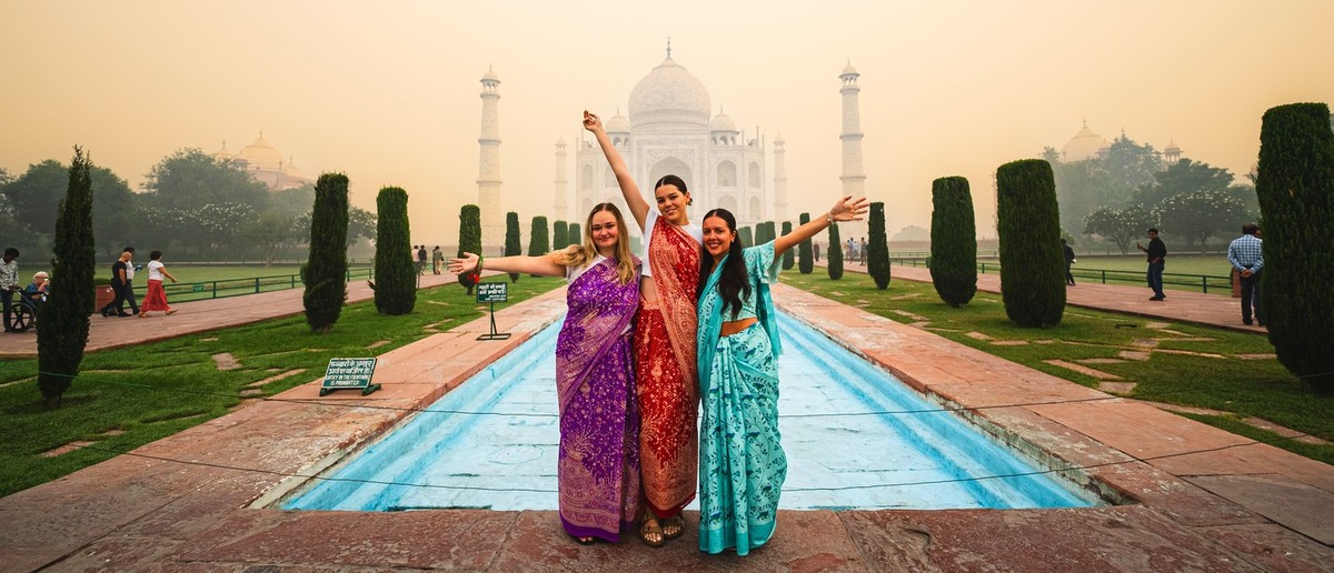 Three women in colourful saris pose with outstretched arms before a majestic white domed building and reflecting pool.