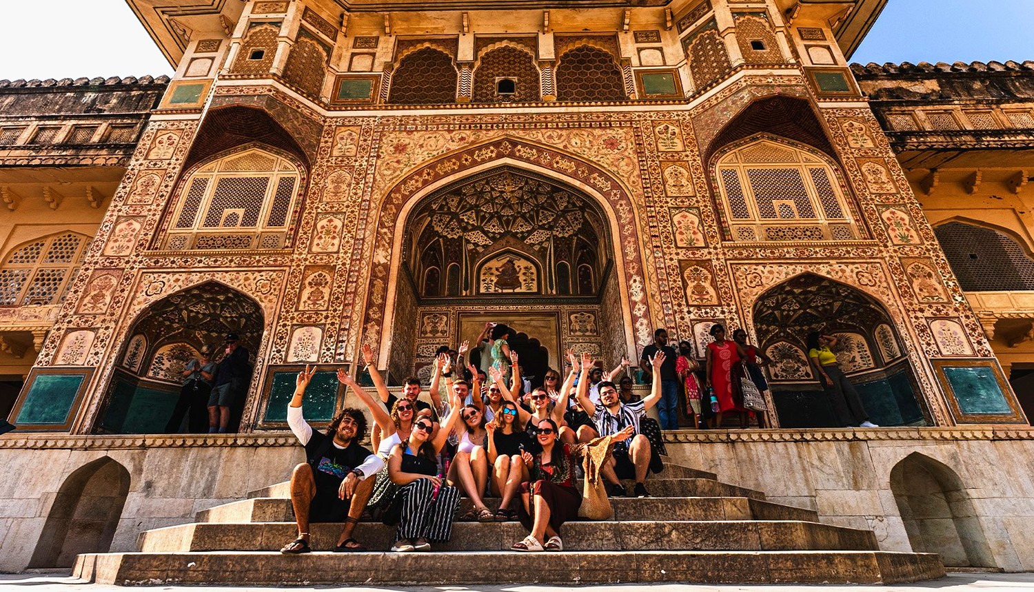 A group of travellers sits on stone steps, smiling and waving in front of an ornately decorated building.