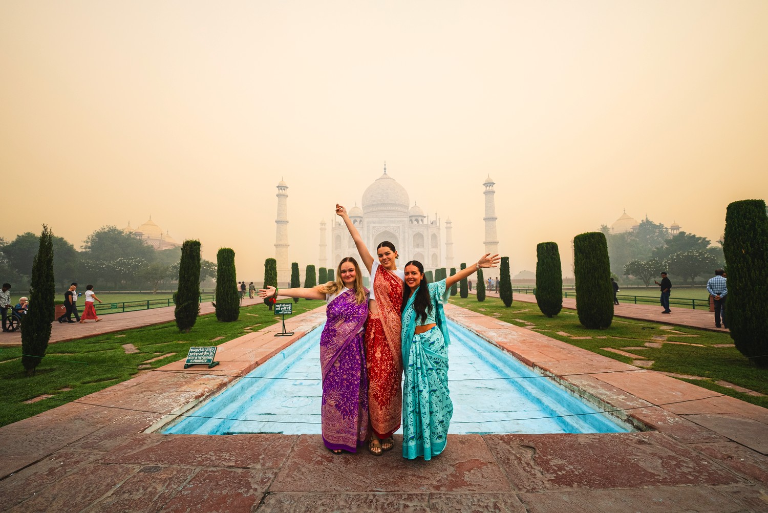 Three women in colourful saris pose with outstretched arms before a majestic white domed building and reflecting pool.