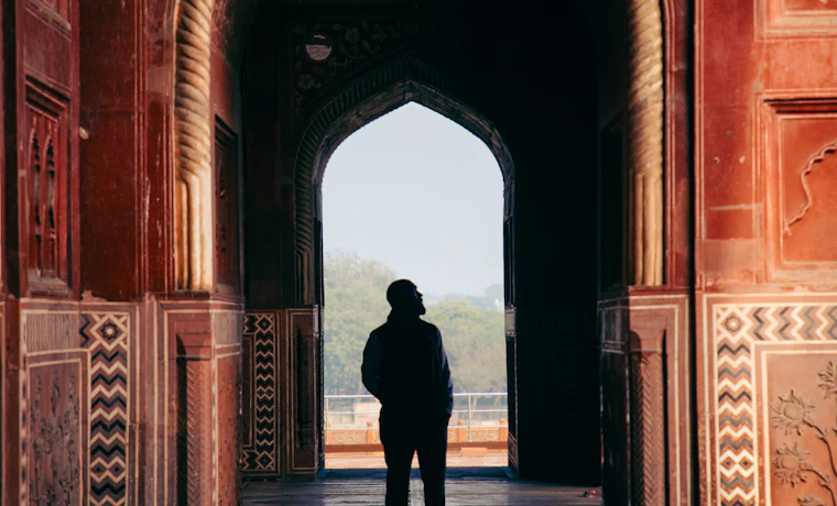 A person stands silhouetted in an ornate red archway, gazing out at the hazy view in Agra.