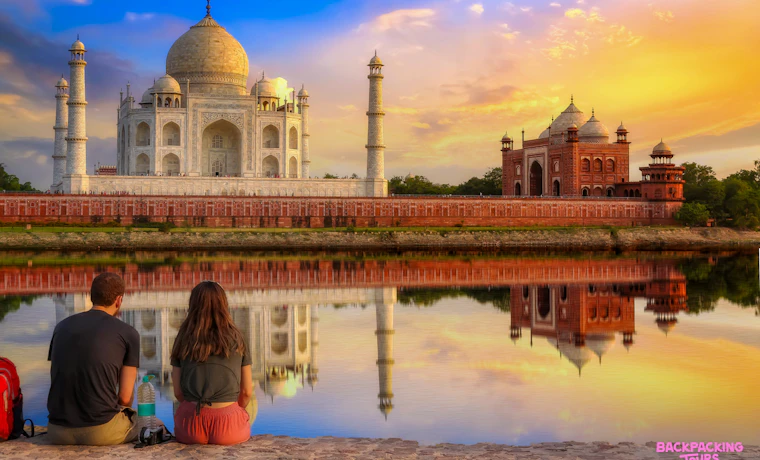 A couple watches the stunning Taj Mahal and its reflection under a vibrant sunrise in Agra.