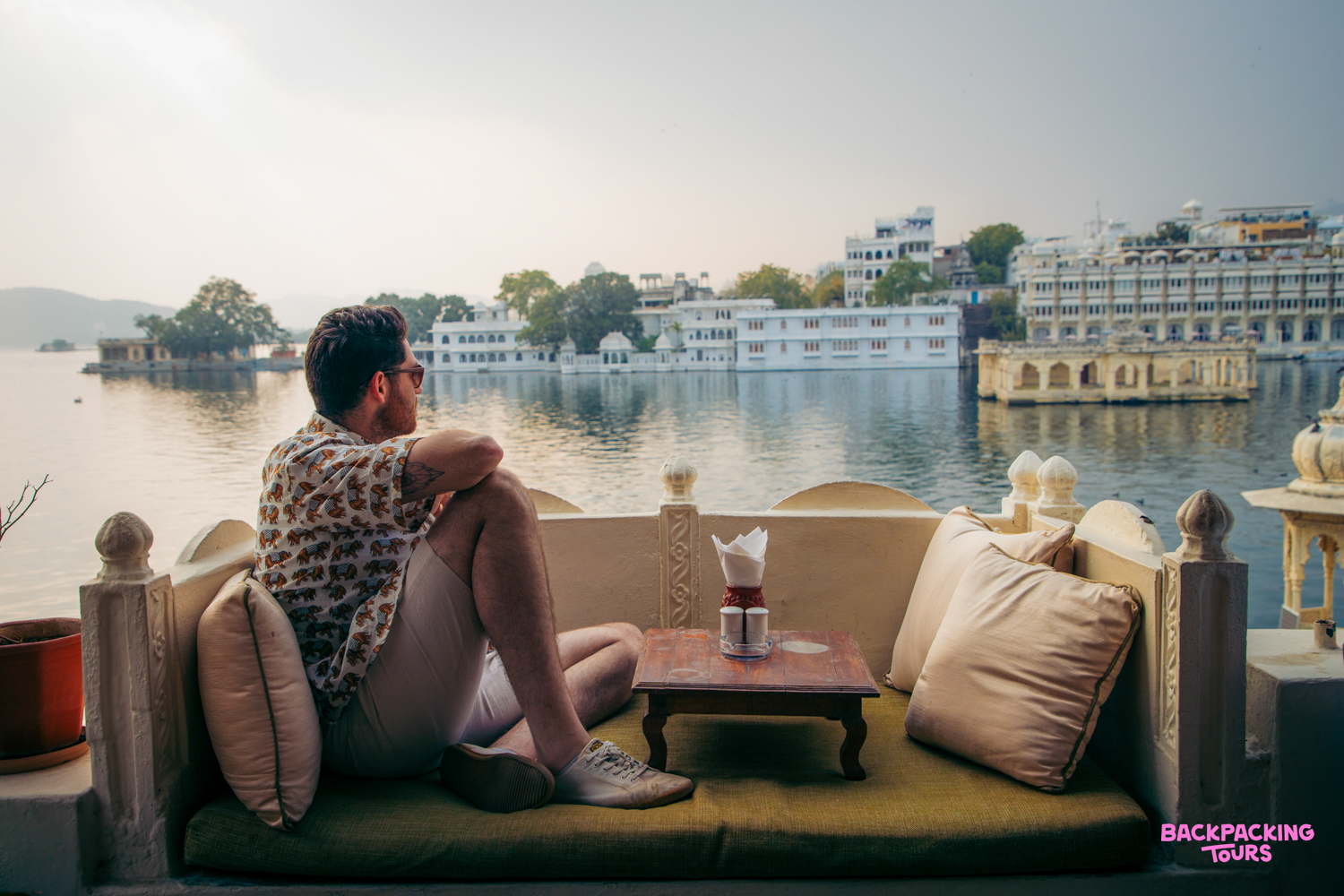A man relaxes on a balcony, admiring the serene waters and grand palaces of Lake Pichola, Udaipur.