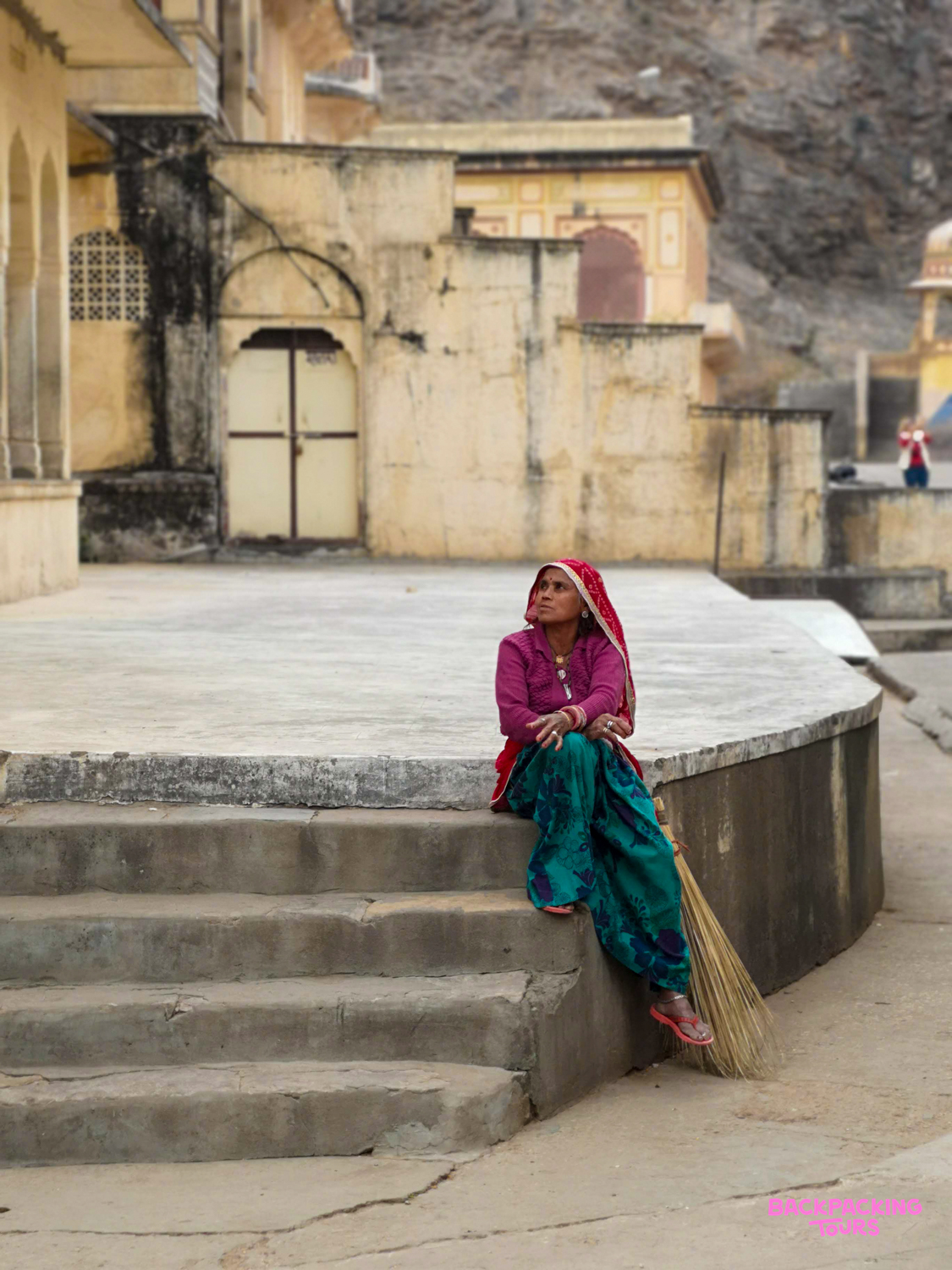 A woman in vibrant traditional attire sits on stone steps with a broom, looking up in Udaipur.