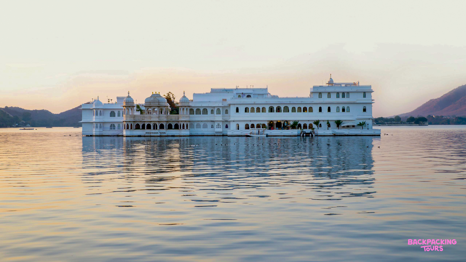 A majestic white palace reflecting on the calm waters of Lake Pichola in Udaipur at sunset.