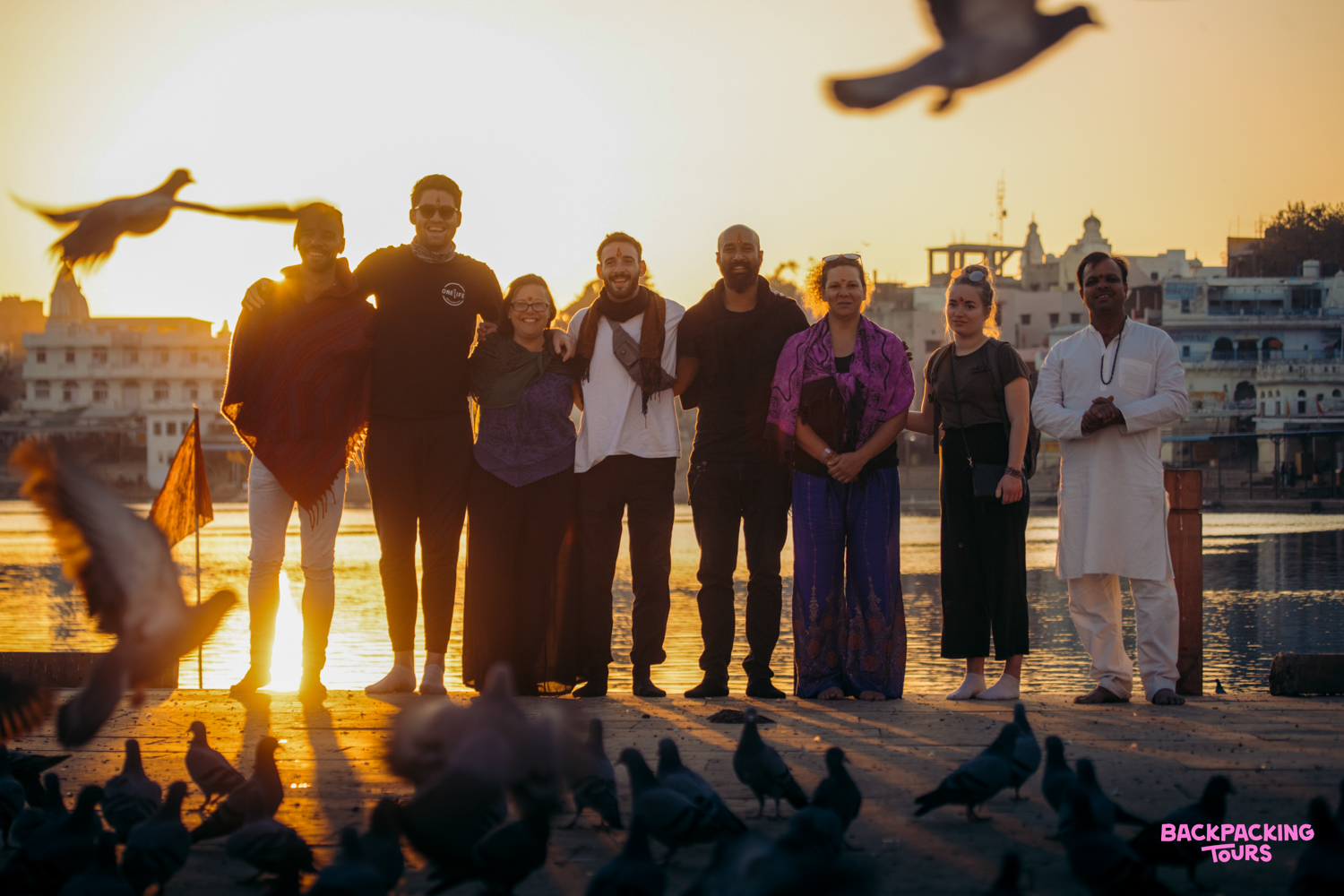 Travelers and a local stand by the sacred Pushkar Lake at sunset, with many pigeons.