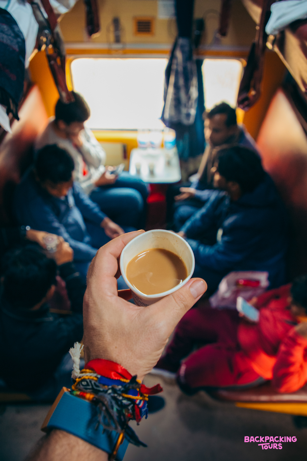 A backpacker enjoys a cup of chai while traveling by train through India with fellow passengers.