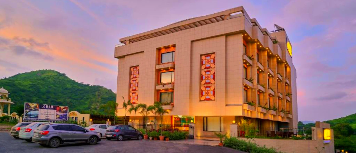 A modern hotel building in Udaipur at sunset, with cars parked and green hills in the background.