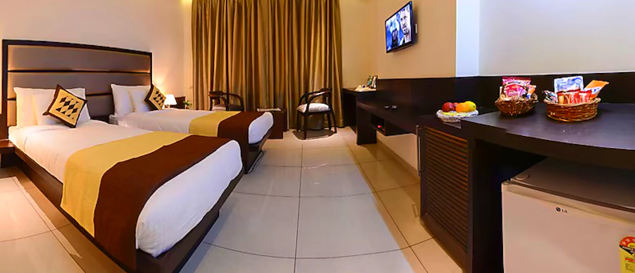 Bright hotel room with two single beds, a desk, mini-fridge, and a TV on the wall.