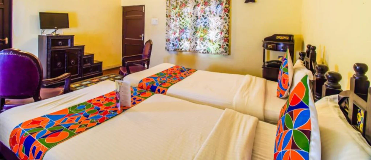 A bright hotel room with two beds, colorful bed runners, and patterned pillows.