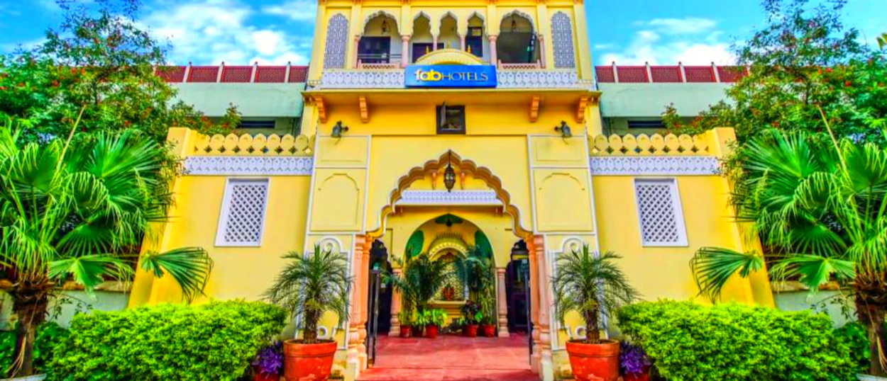 A grand yellow hotel building with traditional Indian architecture and lush green plants in Jaipur.