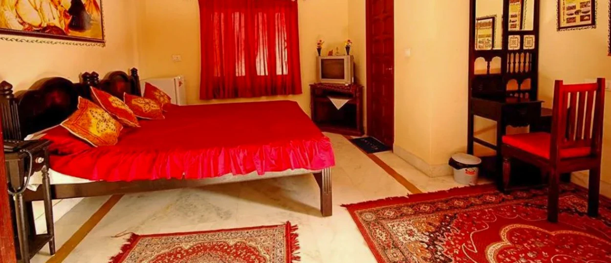 A cozy traditional haveli bedroom in Jodhpur with a red bed, patterned rugs, and wooden furniture.