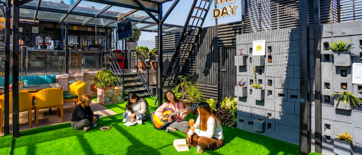 Four friends relax on a vibrant rooftop cafe in Kathmandu, one playing a guitar.