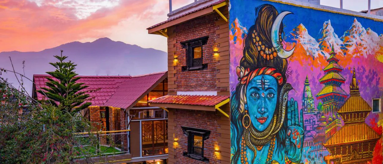 Vibrant mural of blue Shiva and traditional Nepali pagodas on a building, with a sunset over Kathmandu mountains.