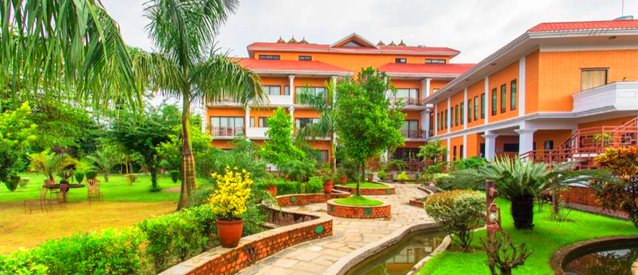 A serene orange lodge with a lush green garden, winding path, and pond in Chitwan National Park.