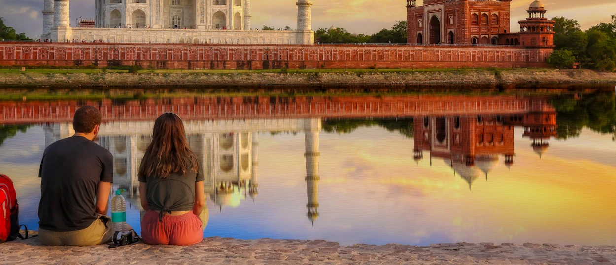 A couple sits by the water, gazing at the Taj Mahal and its reflection during sunrise in Agra.