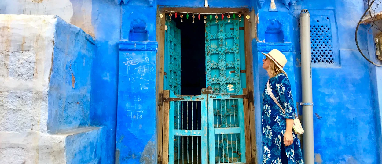 A traveler in a blue dress and hat stands by a vibrant blue wall and ornate door in Jodhpur.