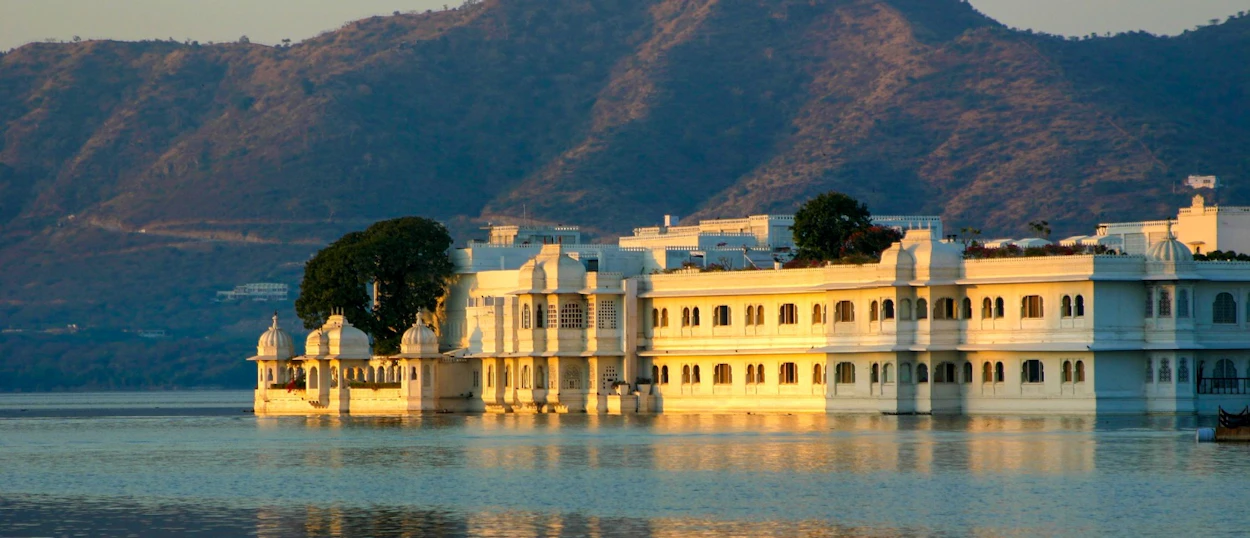 Elegant white palace reflecting in the calm waters of Lake Pichola at sunset, with distant mountains.