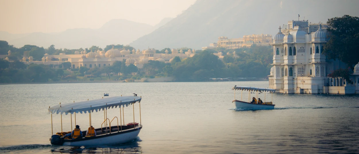 Two boats cruise on Lake Pichola with historic white palaces and hills in Udaipur.