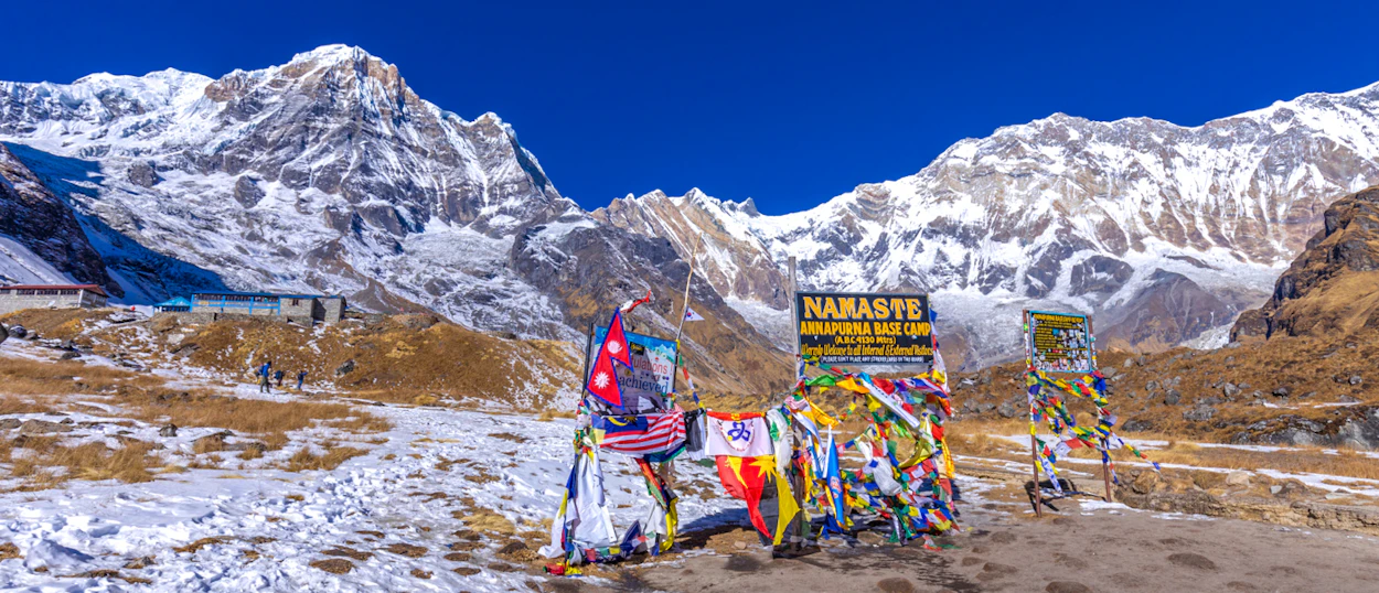 Colorful prayer flags mark the snowy Annapurna Base Camp with majestic mountains under a clear blue sky.