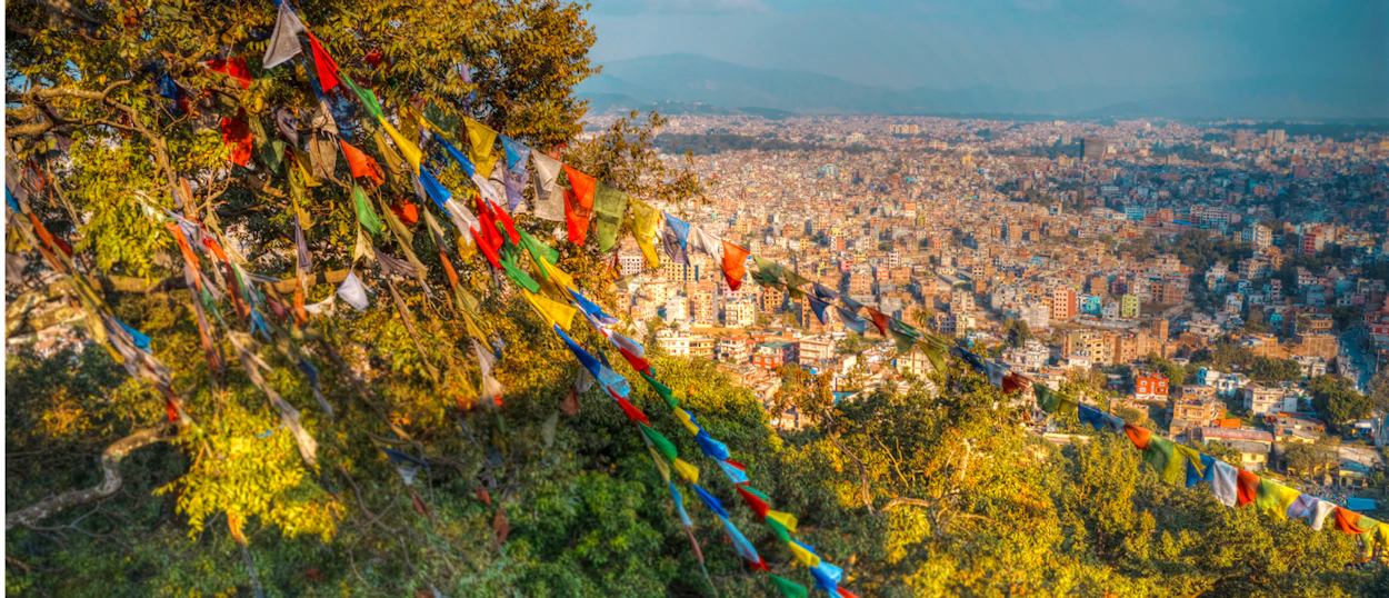 Colorful prayer flags wave over a sprawling city view of Kathmandu at sunset.