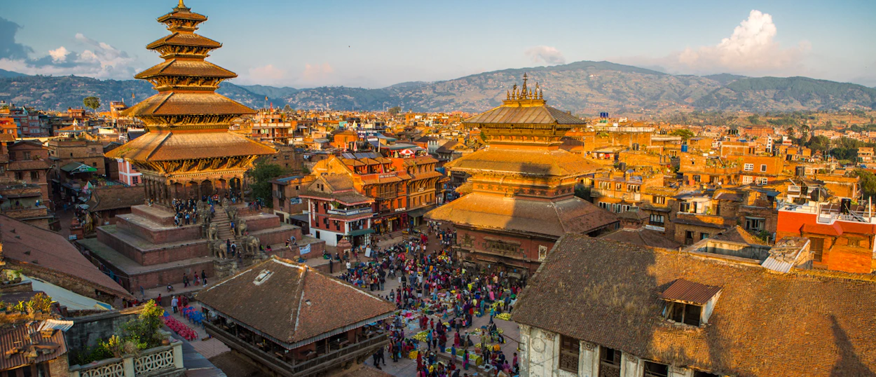 Crowds gather in Kathmandu's historic Durbar Square, surrounded by ornate tiered temples and distant mountains.