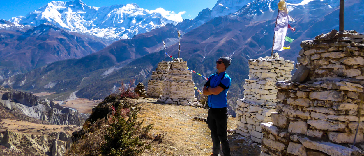 A hiker stands on a ridge near stone stupas, overlooking the vast Annapurna mountain range.