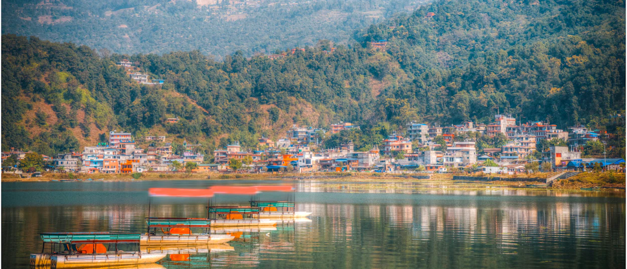 Colorful boats float on serene Phewa Lake, reflecting the vibrant town of Pokhara and surrounding green hills.