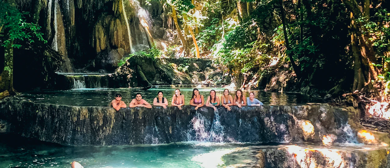 Nine women relax in clear natural pools at the base of lush waterfalls on Moyo Island.
