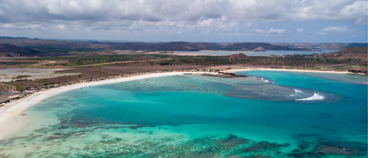 Aerial view of a pristine white sand beach and clear turquoise ocean water with distant hills in Lombok.