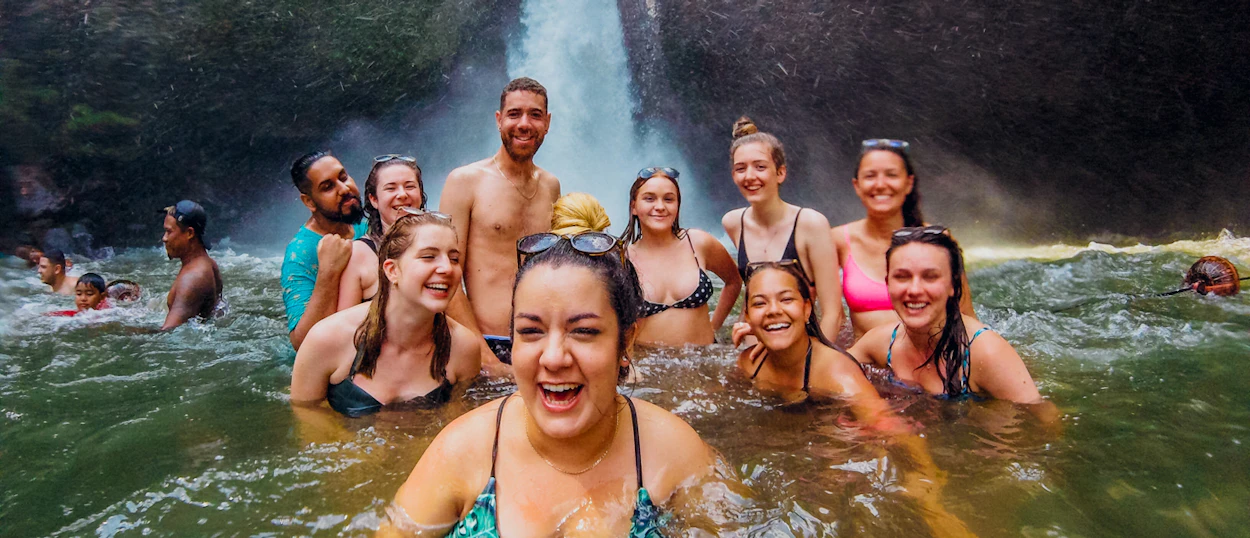 A joyful group of backpackers swimming and laughing at the base of a waterfall in Ubud, Bali.
