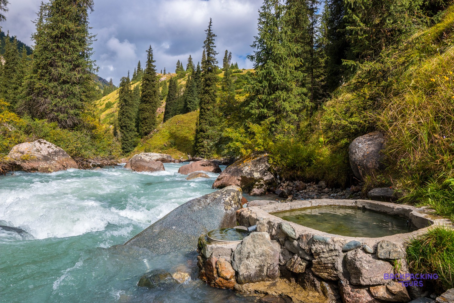 A stone-lined hot spring pool sits beside a rushing river, surrounded by green hills and tall evergreen trees.