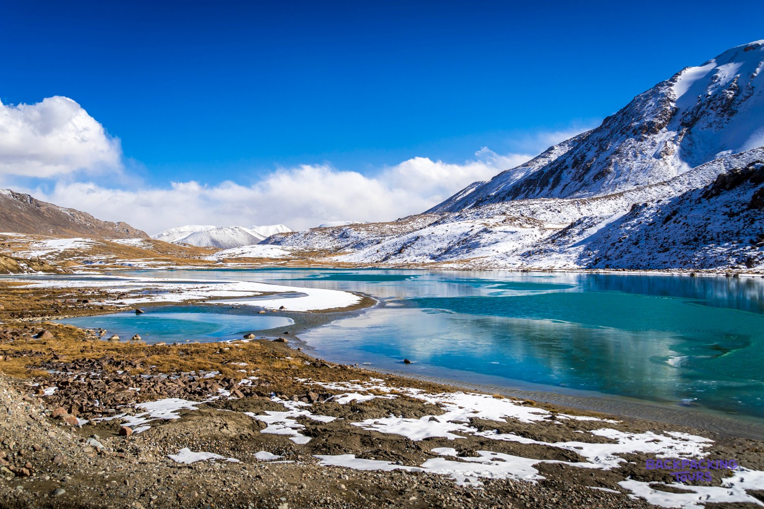 Turquoise lake surrounded by snow-dusted mountains under a bright blue sky with white clouds.