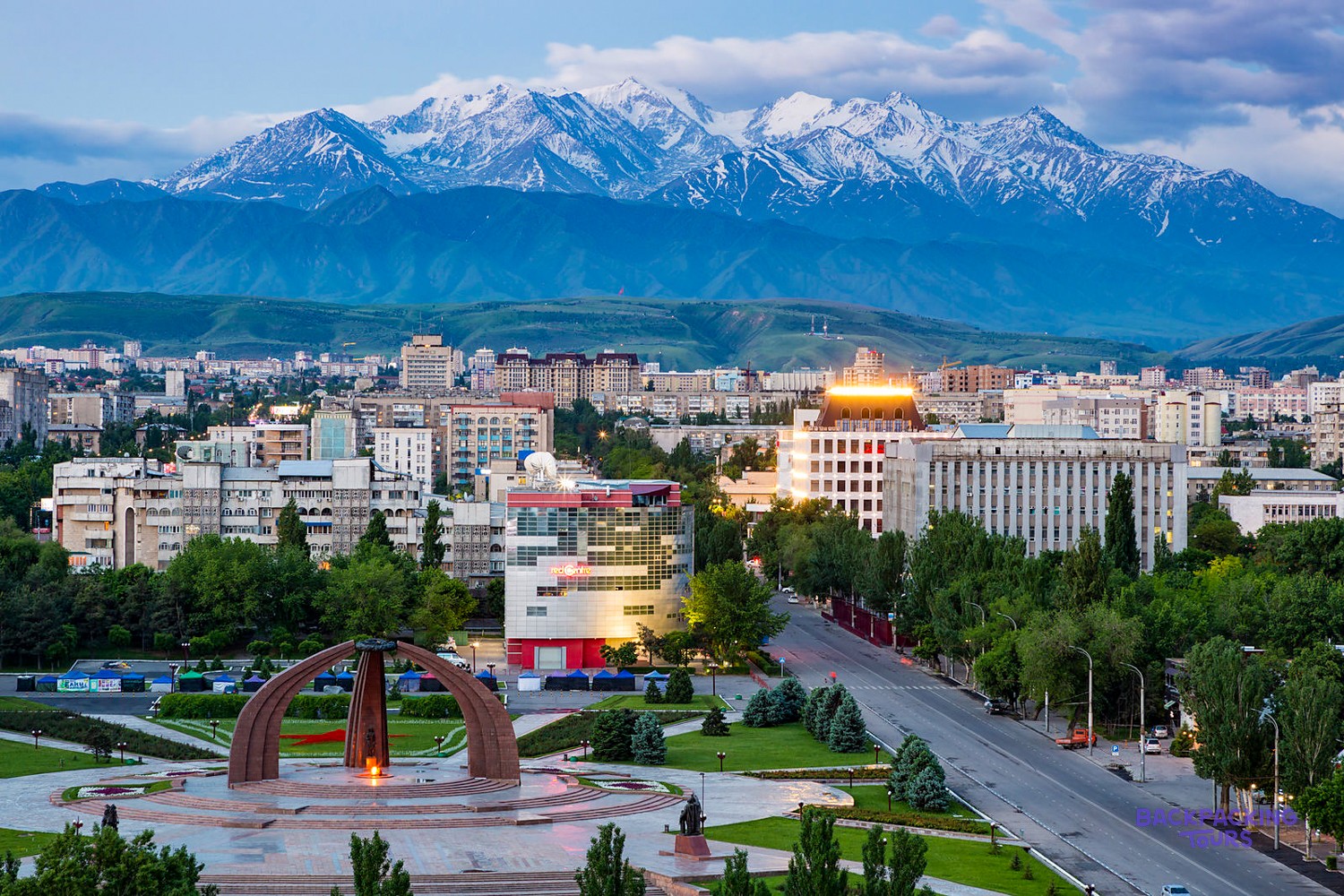 A city skyline with numerous buildings and green trees is set against a backdrop of towering, snow-capped mountains.