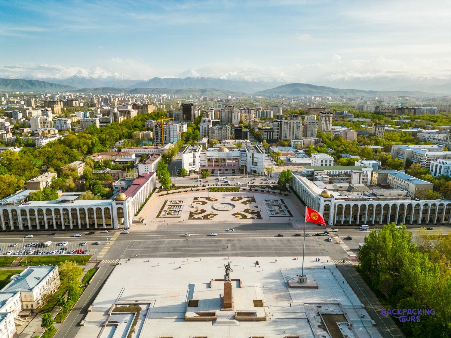 An expansive city square features a statue, a red flag, buildings, and distant snow-capped mountains.