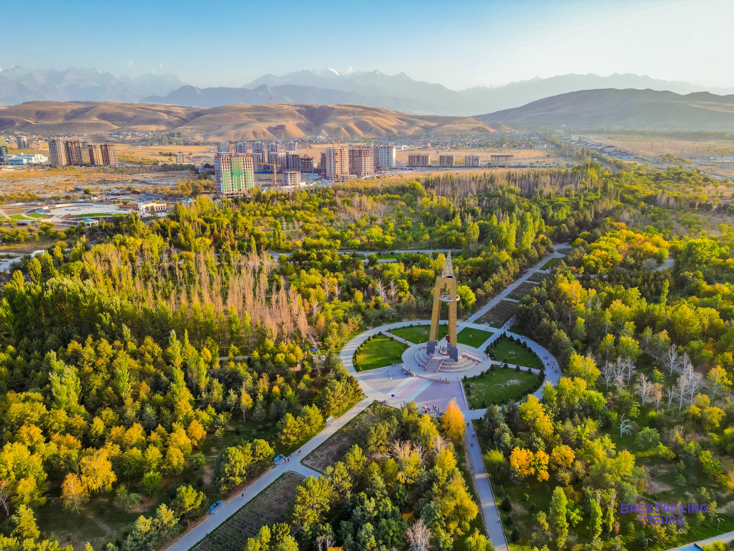 A large park with a golden monument, winding paths, and autumn trees, backed by buildings and mountains.