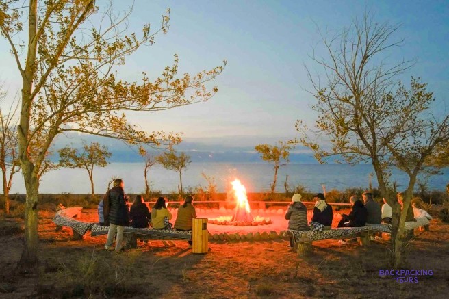 A group of people sit on benches around a large bonfire by a body of water at dusk.