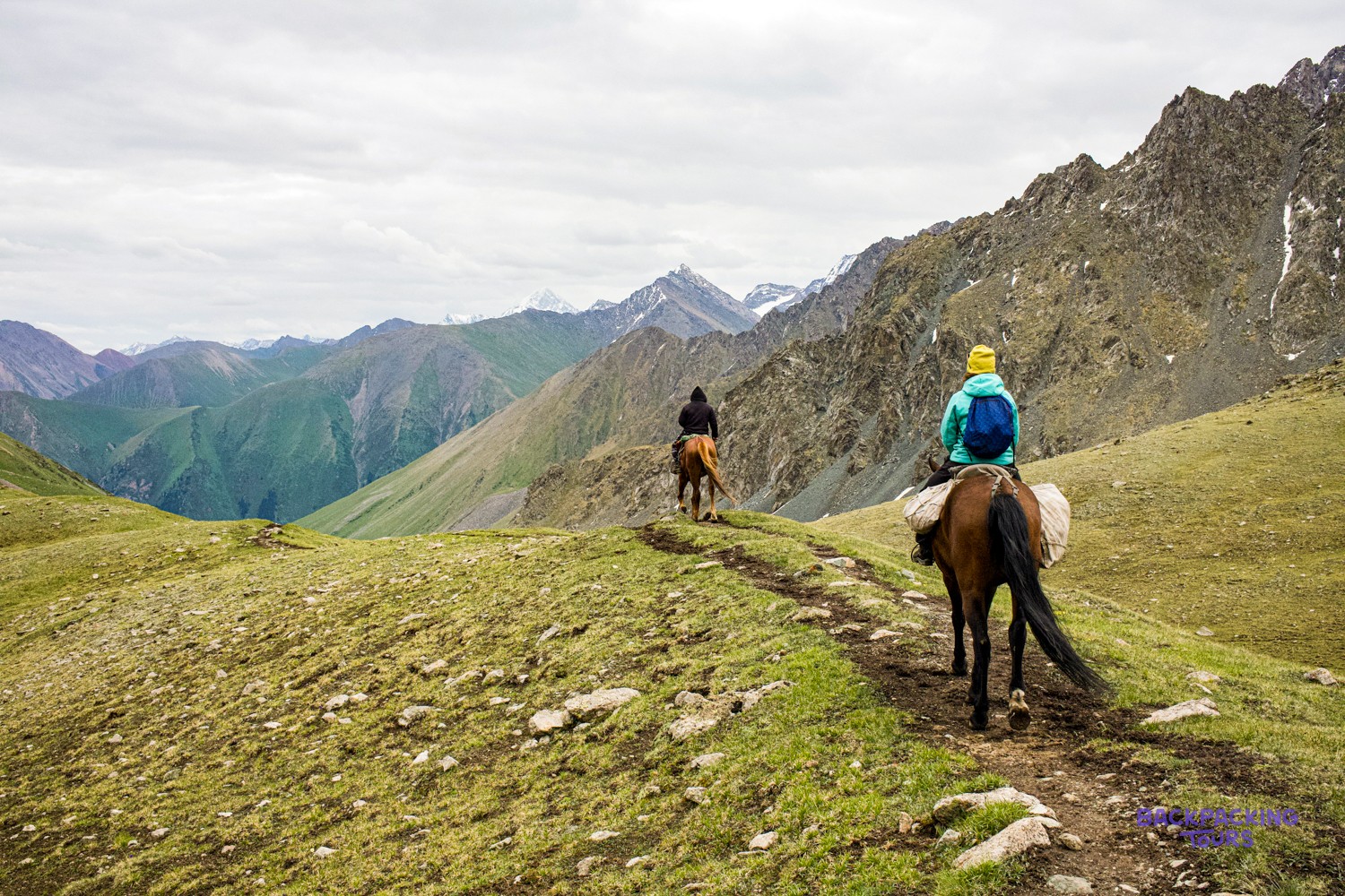 Two riders on horseback follow a trail through vast, rocky mountains with green slopes and distant peaks.