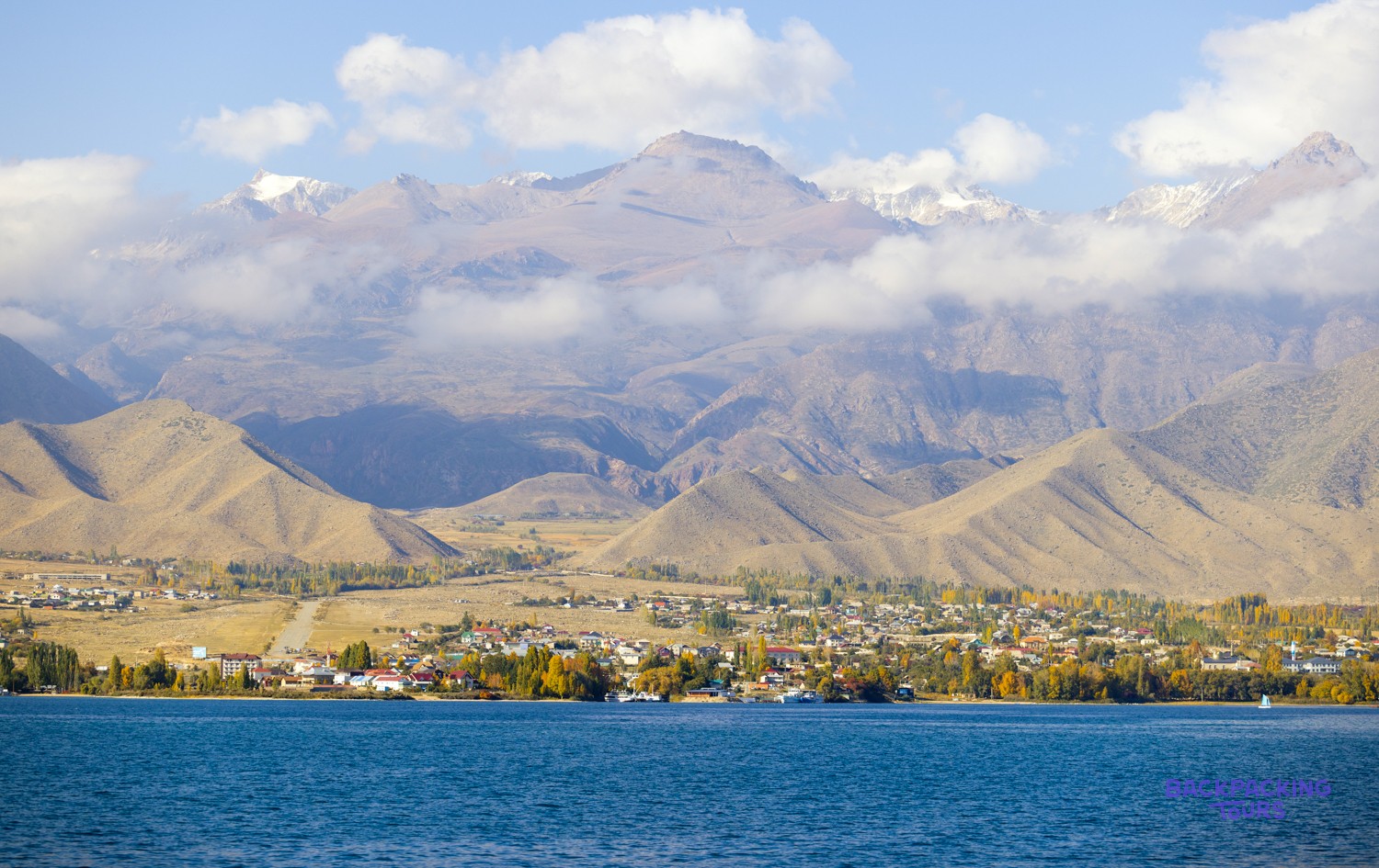A town with colourful autumn trees lines a blue lake, backed by cloud-covered, snow-capped mountains.