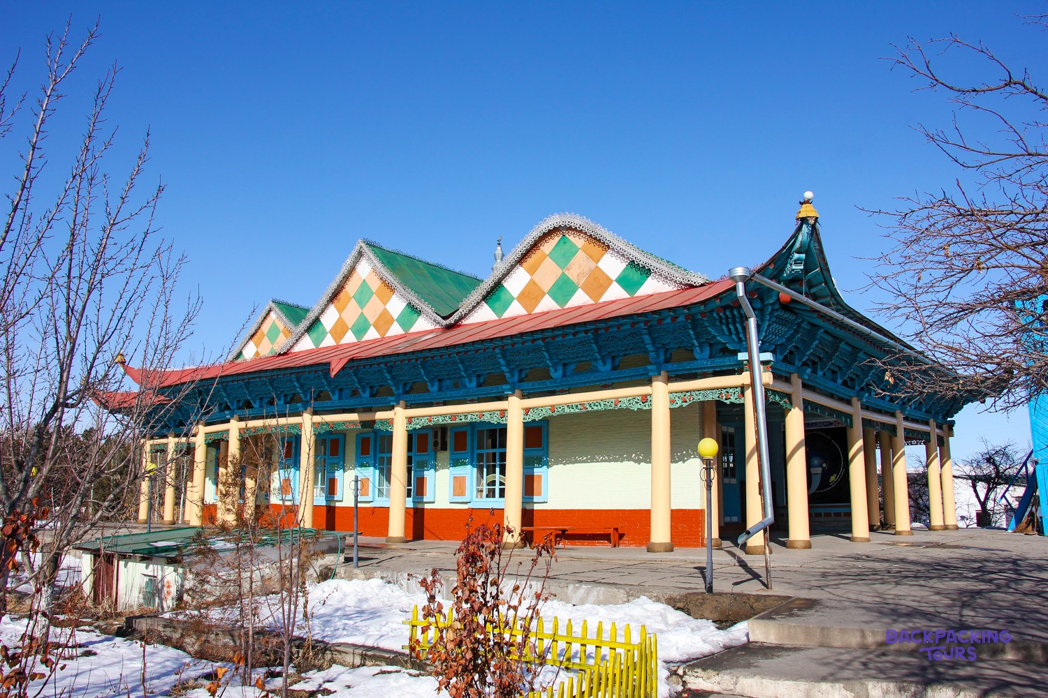A colourful building with ornate blue eaves, yellow columns, and a green and orange diamond-patterned roof stands under a clear sky.