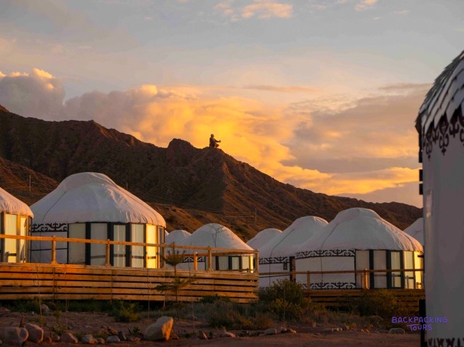 Several white yurts with wooden decks are nestled at the base of mountains under a vibrant sunset.