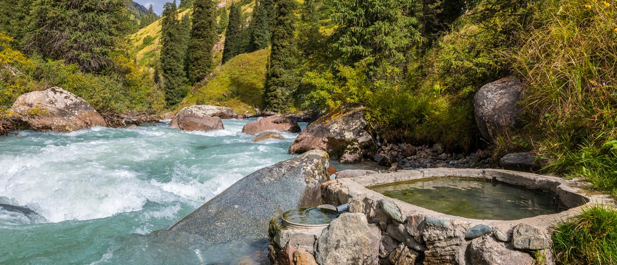 A stone-lined hot spring pool sits beside a rushing turquoise river in a green, forested valley.