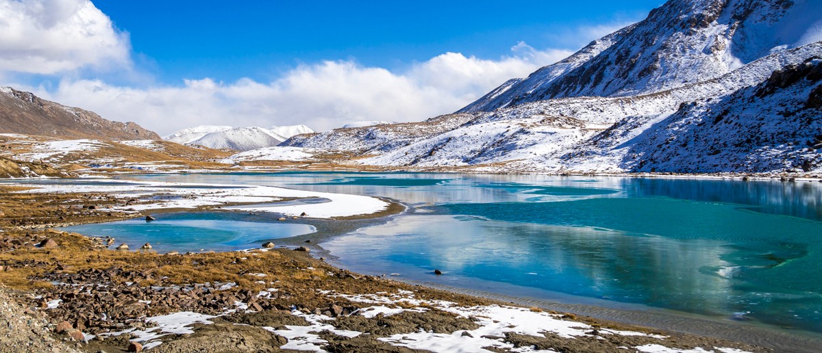 A bright blue lake surrounded by snow-dusted mountains and rocky, grassy terrain under a blue sky.
