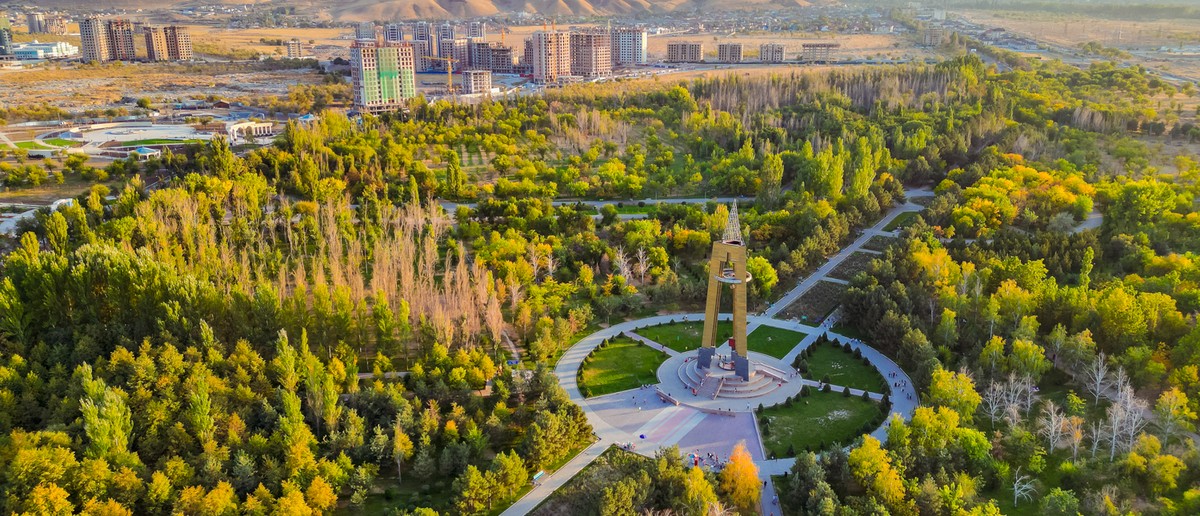A large golden monument in a green park with circular paths, surrounded by city buildings and distant hills.