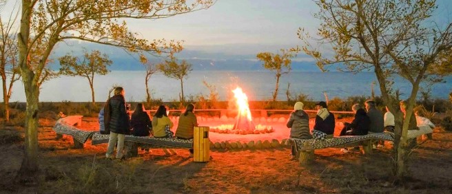 A group of people sit around a large bonfire on benches near a body of water at dusk.