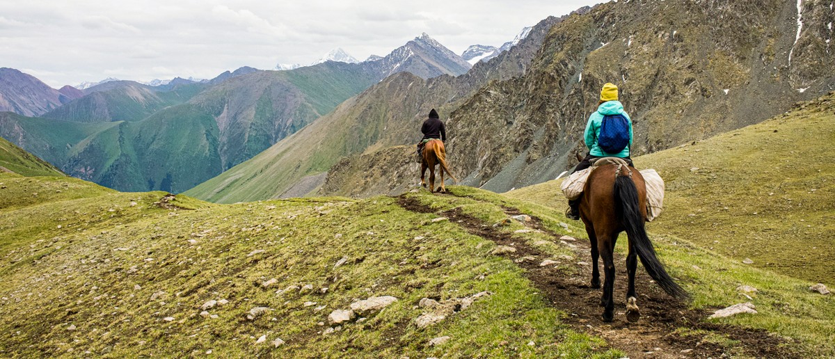 Two people on horseback ride a winding trail through green, rocky mountains under a cloudy sky.