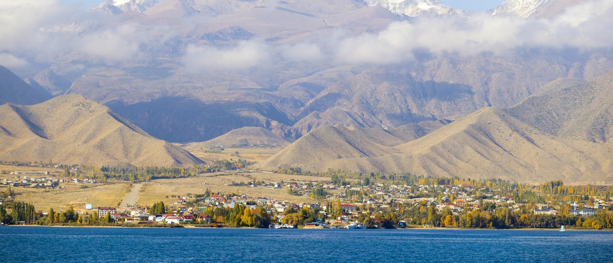 A town with colourful trees sits by a blue lake, backed by dry hills and snow-capped mountains.