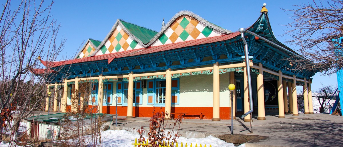 A long, ornate building with green and orange diamond roofs, blue carved eaves, and yellow pillars.